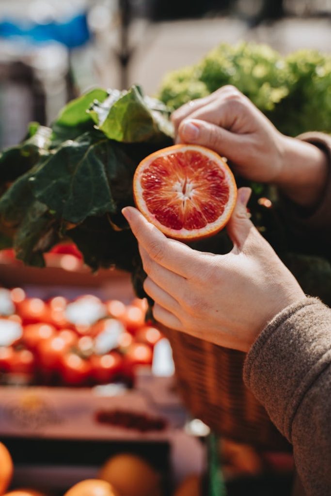 pexels-photo-7342216 Close-up of hands holding a sliced orange with vegetables in a market setting, showcasing freshness and vibrancy.